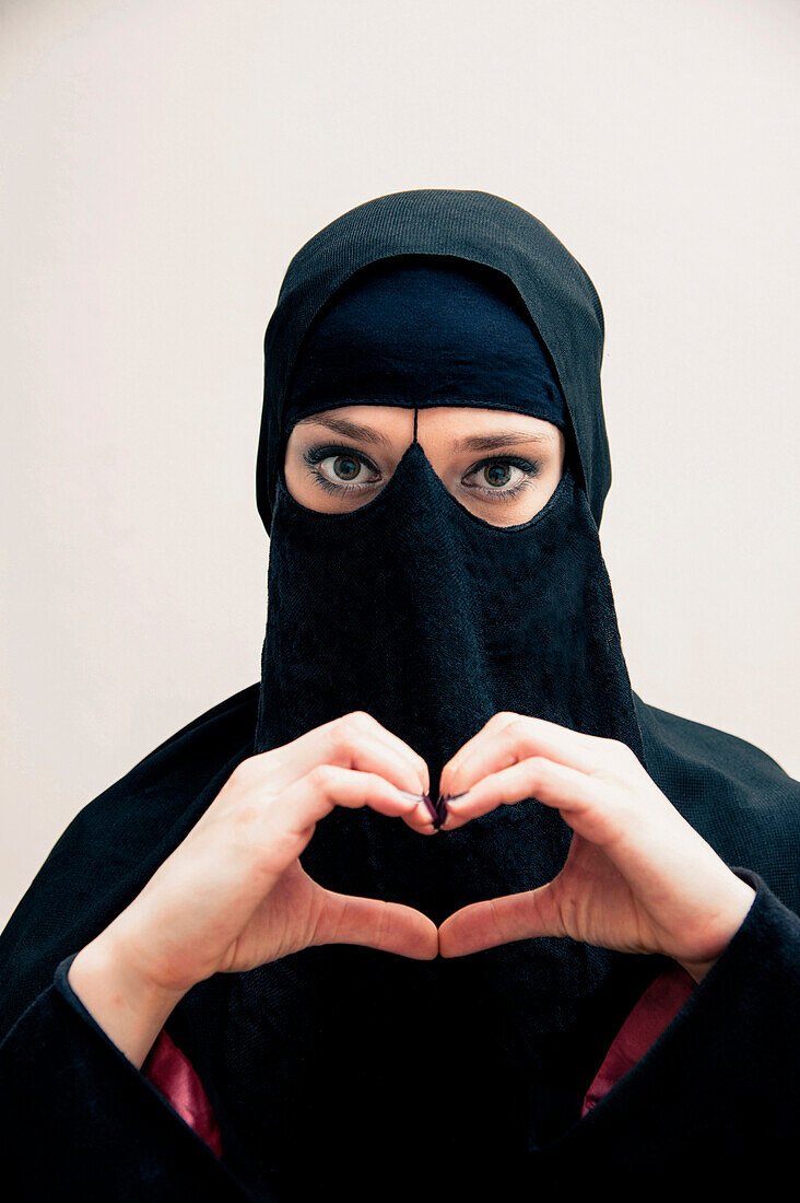 Nahaufnahme einer jungen Frau in schwarzem muslimischen Hijab und muslimischem Kleid, die mit ihren Händen eine Herzform bildet, in die Kamera schaut und ihre Augen geschminkt hat, Studioaufnahme auf weißem Hintergrund