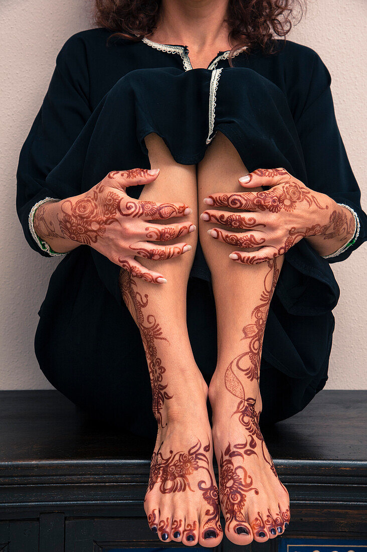 Tiefschnitt einer Frau, die in einem Raum sitzt und Beine, Füße und Hände mit Henna im arabischen Stil bemalt, in einem typischen schwarzen, arabischen, muslimischen Kleid, Studioaufnahme