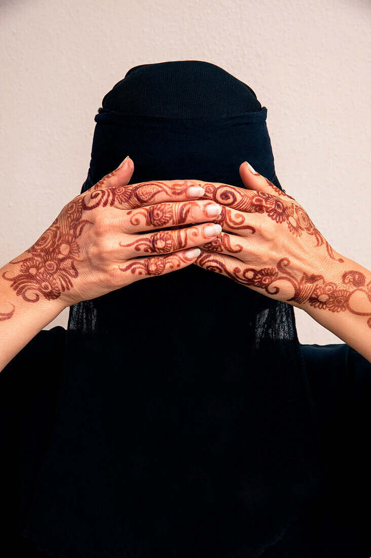 Nahaufnahme einer Frau, die einen schwarzen muslimischen Hidschab und ein muslimisches Kleid trägt, die Hände bedecken die Augen und zeigen mit Henna im arabischen Stil bemalte Hände, Studioaufnahme vor weißem Hintergrund