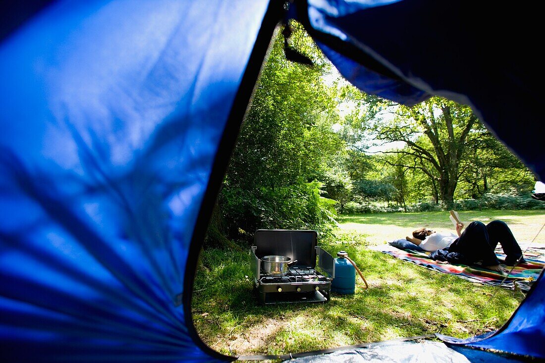 Blick durch ein Zelt auf einen Campingplatz und eine lesende Frau