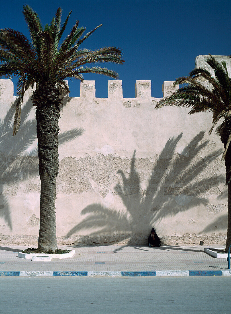 Woman In Djelleba Resting In Shadow Of Date Palm Against Town Walls