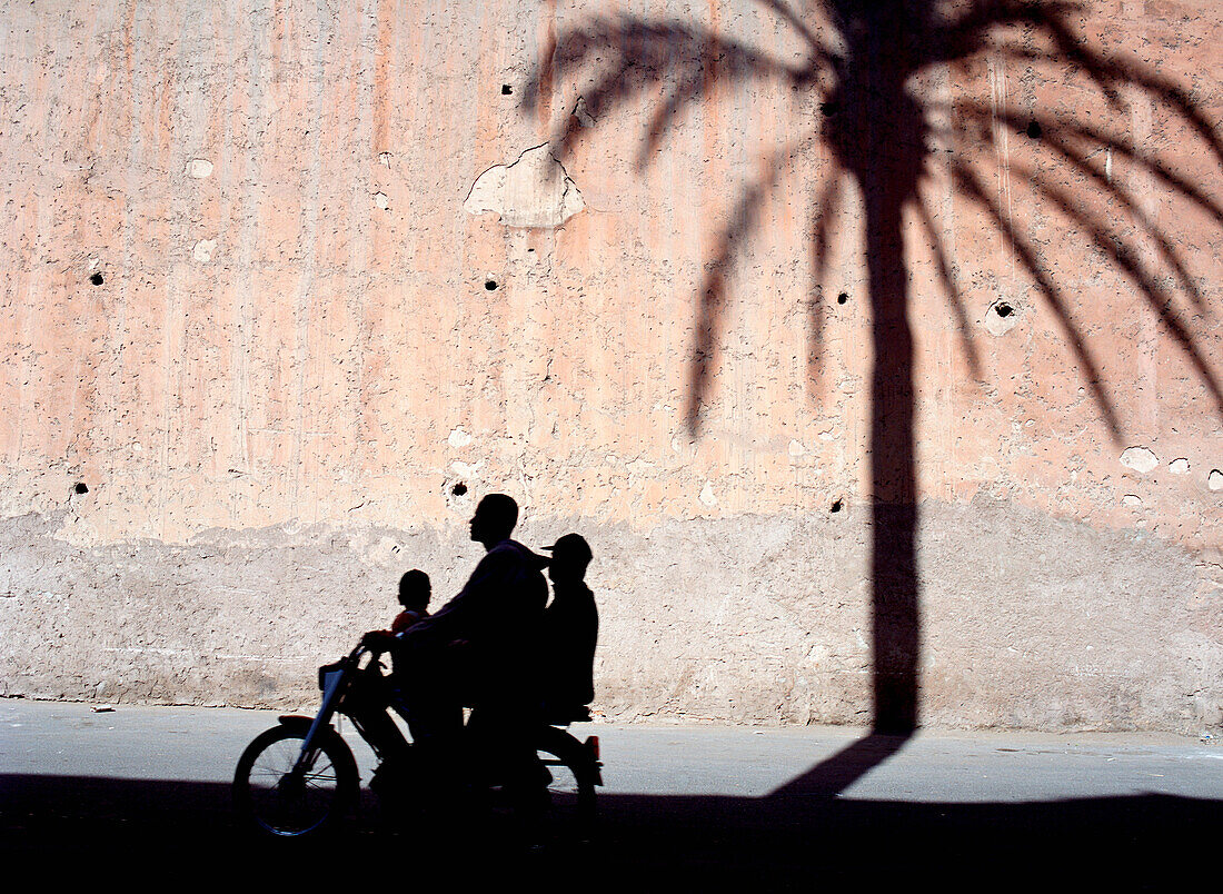 Silhouette eines Vaters und zweier Söhne auf einem Moped, die am Schatten einer Dattelpalme an der Stadtmauer vorbeifahren
