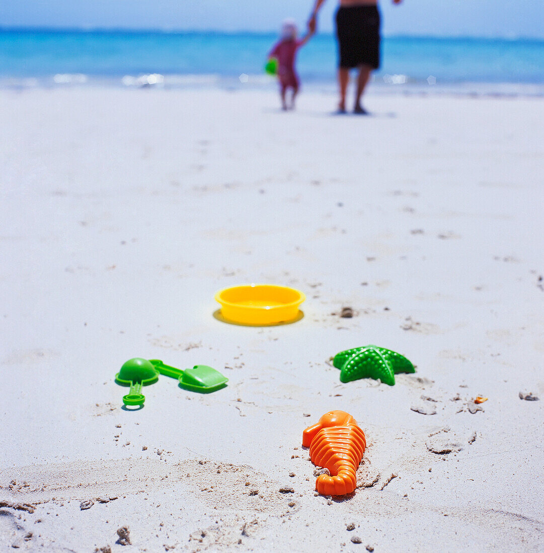 Vater und Tochter gehen am Strand spazieren, im Vordergrund Plastikspielzeug