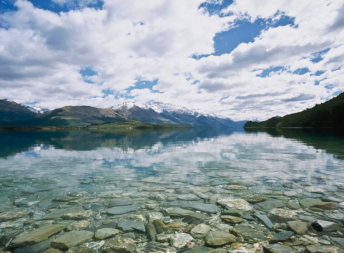 Schneebedeckte Berge spiegeln sich im Lake Wakatipu mit klarem Wasser und Kieselsteinen