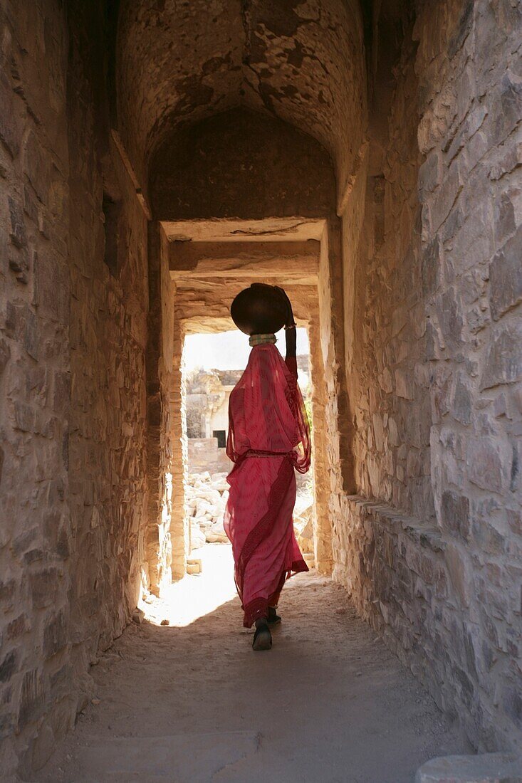 Woman Walking Between Two Stock Buildings Carrying A Pot On Her Head