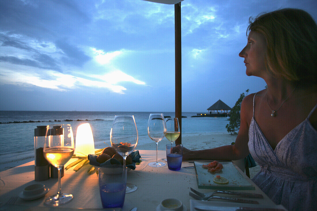 Frau am Esstisch mit Blick auf den Strand in der Abenddämmerung.