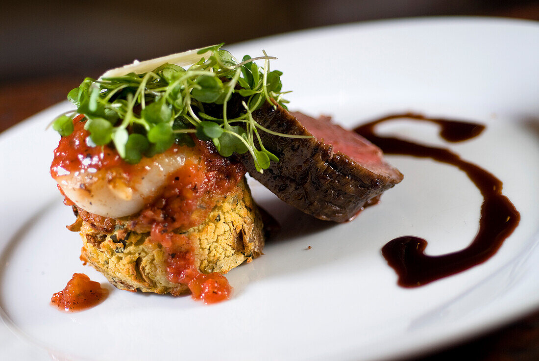 Kobe Beef Tenderloin gebraten und in Scheiben geschnitten, begleitet von bronzierter Alaskan Weathervane Scallop und serviert auf einem Yukon Gold Kartoffelkuchen mit frischem Baby Rucola, gehobeltem Asiago Käse, Balsamico Kalbsreduktion und gerösteten Tomatenkonserven