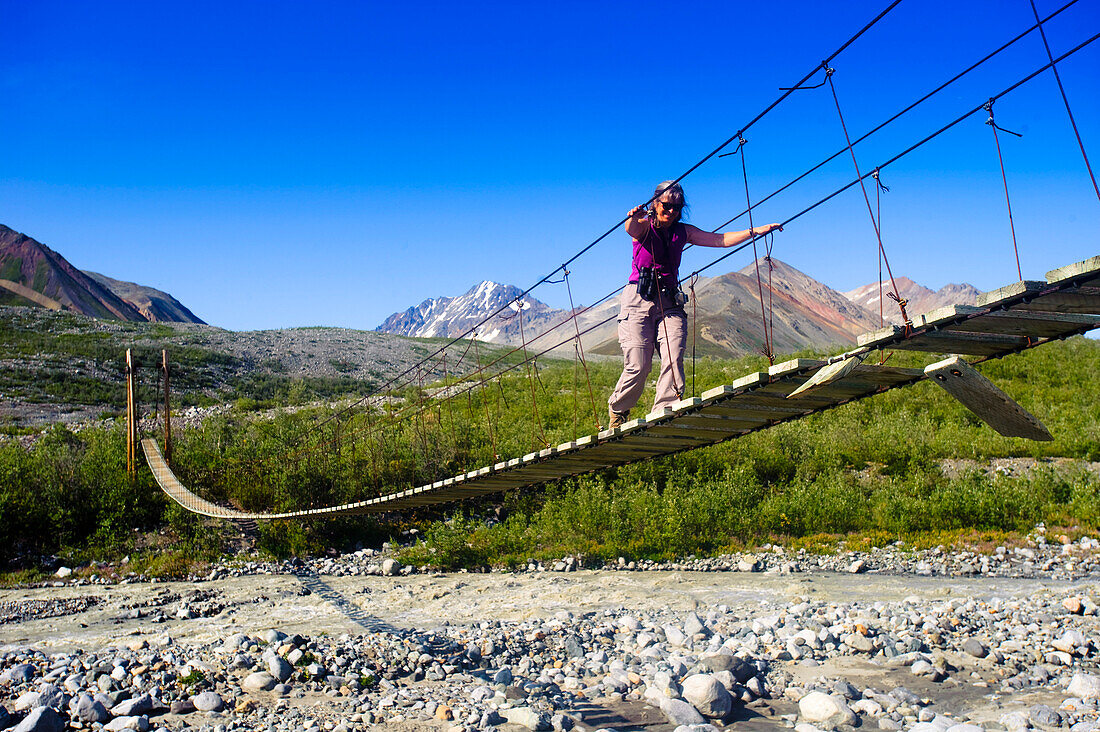 Eine Frau wandert auf einer Hängebrücke über den College Creek zum Gulkana Gletscher, Süd-Zentral-Alaska, Sommer/N