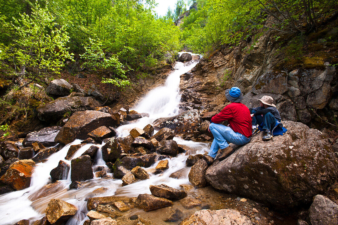 A Father And Son On A Hike Rest On A Rock While Looking At Cascade Falls Near Byers Lake In Denali State Park, Southcentral Alaska, Summer