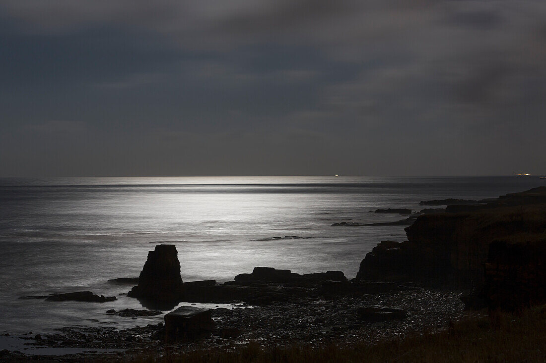 Sonnenlicht spiegelt sich auf dem ruhigen Wasser in der Abenddämmerung mit einer silhouettierten Küstenlinie; South Shields, Tyne And Wear, England