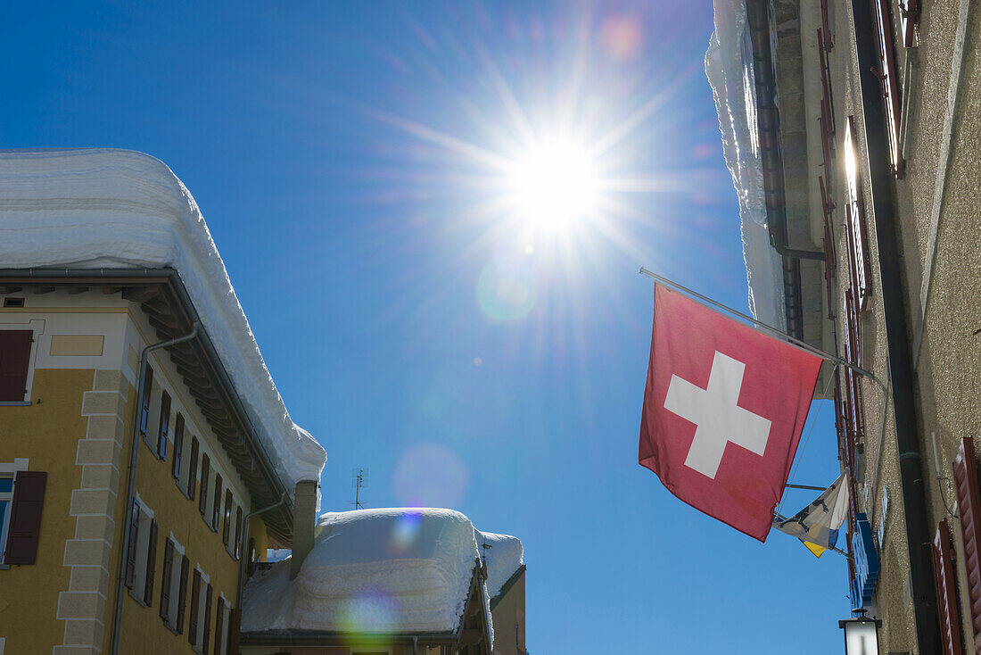 Flag Of Switzerland Hanging From A Building With A Sunburst In A Blue Sky; San Bernardino, Grisons, Switzerland