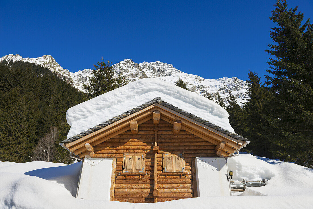 Schneebedeckte Blockhütte mit einer Bergkette im Hintergrund; San Bernardino, Graubünden, Schweiz
