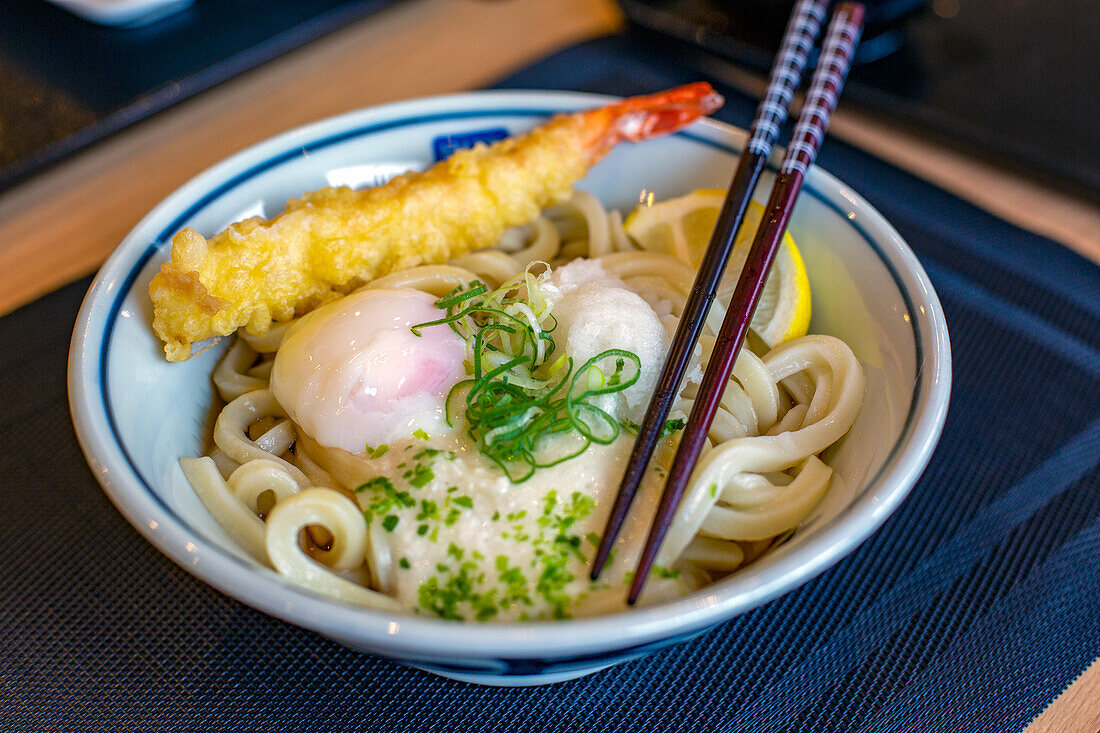 Ein Gericht aus Nudeln, Gemüse und Fisch sowie eine Tempura-Garnele, Stäbchen. 