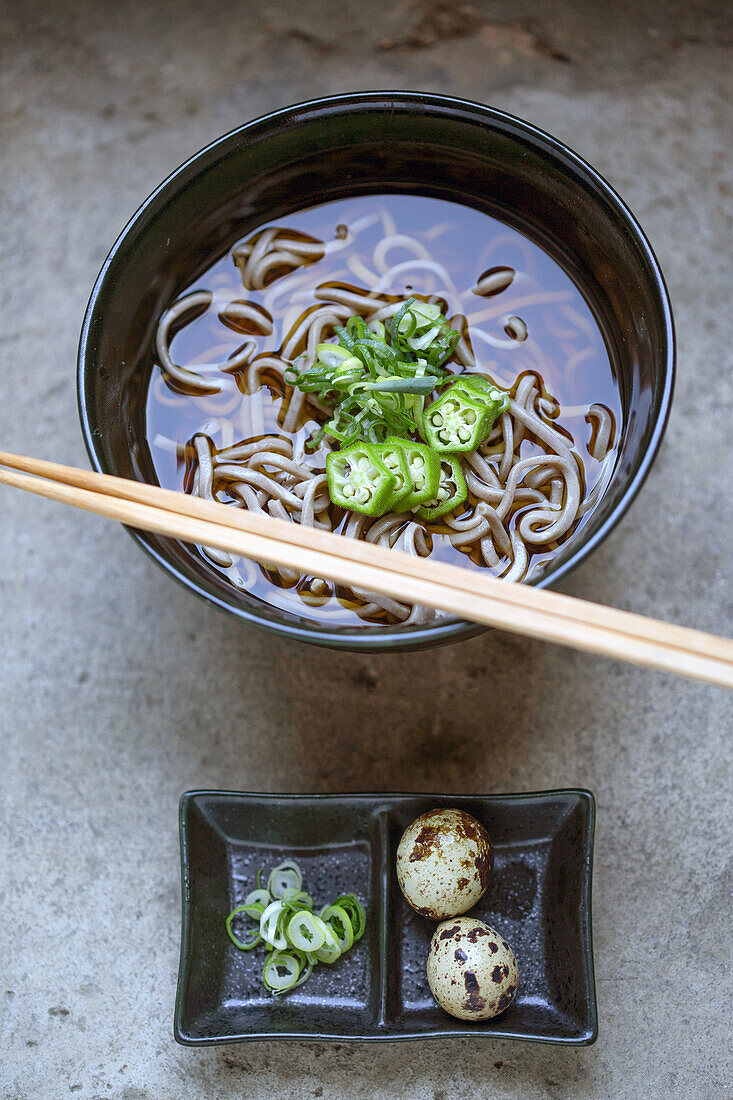 A black china bowl of noodles and broth, sliced green chilli and a dish with quail's eggs and sliced vegetables.