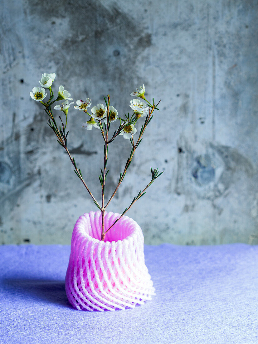 Studioaufnahme, ein Stiel kleiner weißer Blumen in einer rosafarbenen Vase aus recyceltem Plastiknetz. 