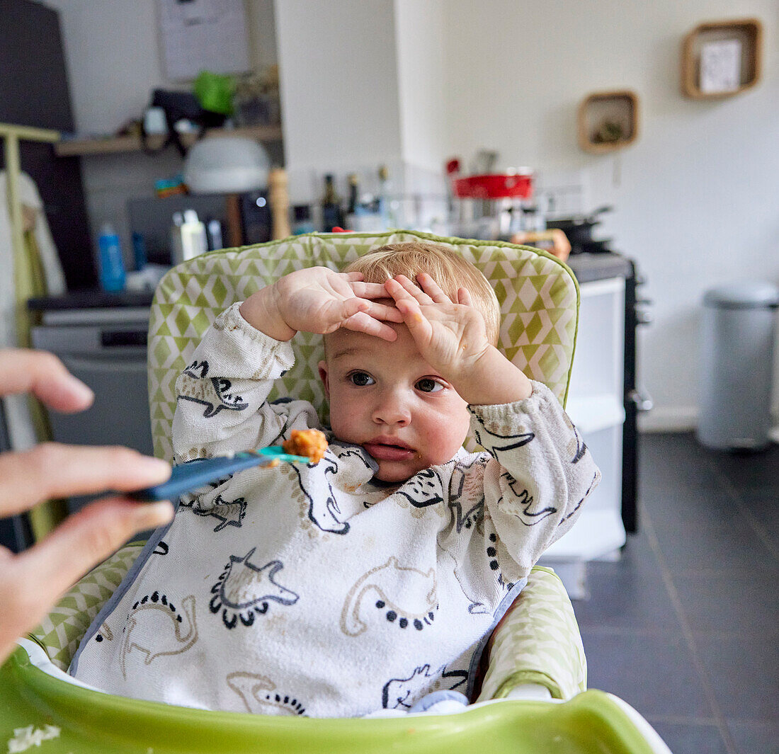 Kleinkind mit erhobenen Händen, das im Hochstuhl in der Küche sitzend das Essen verweigert