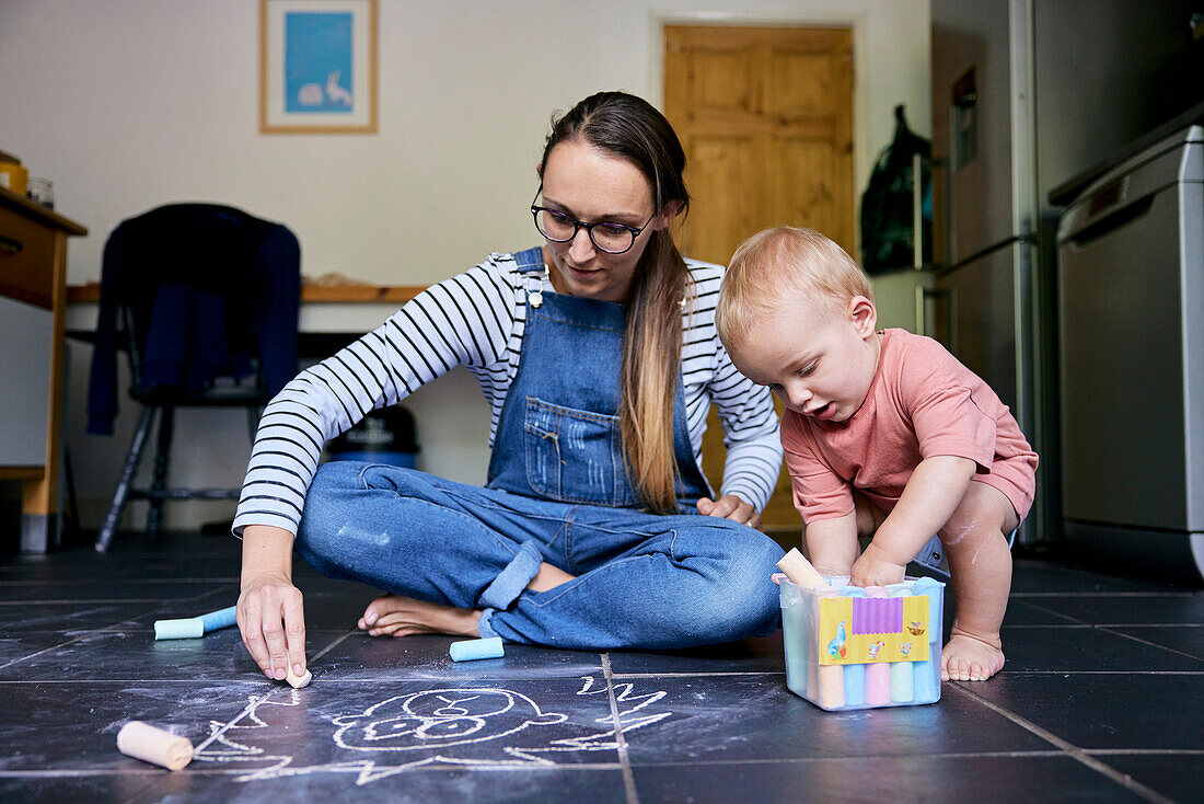 Mother and toddler drawing with chalks together on a tiled kitchen floor