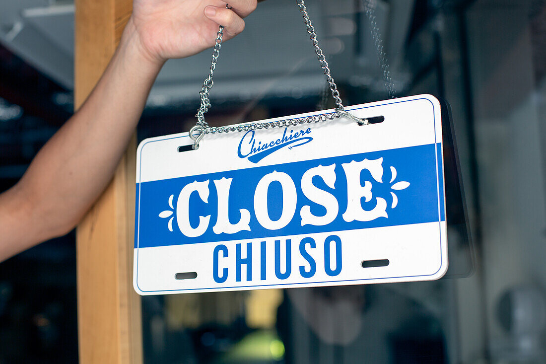 A person hanging up a sign on a restaurant door, reading Close. Closed, dual language, English and Italian.