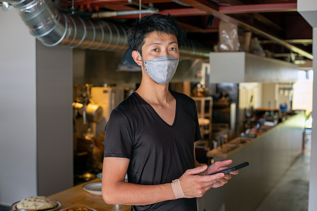 A man working in a restaurant, wearing a face mask, by an open kitchen, holding a digital tablet. 