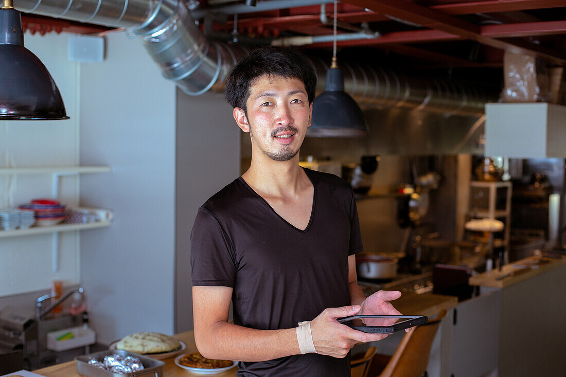 A man working in a restaurant, by an open kitchen, holding a digital tablet. 