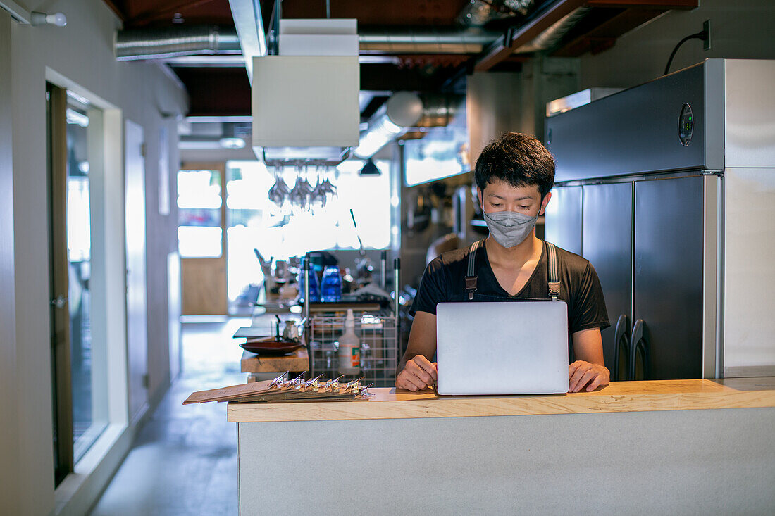 Ein Mann mit einer Gesichtsmaske in einer Restaurantküche, der einen Laptop benutzt, der Besitzer oder Manager. 