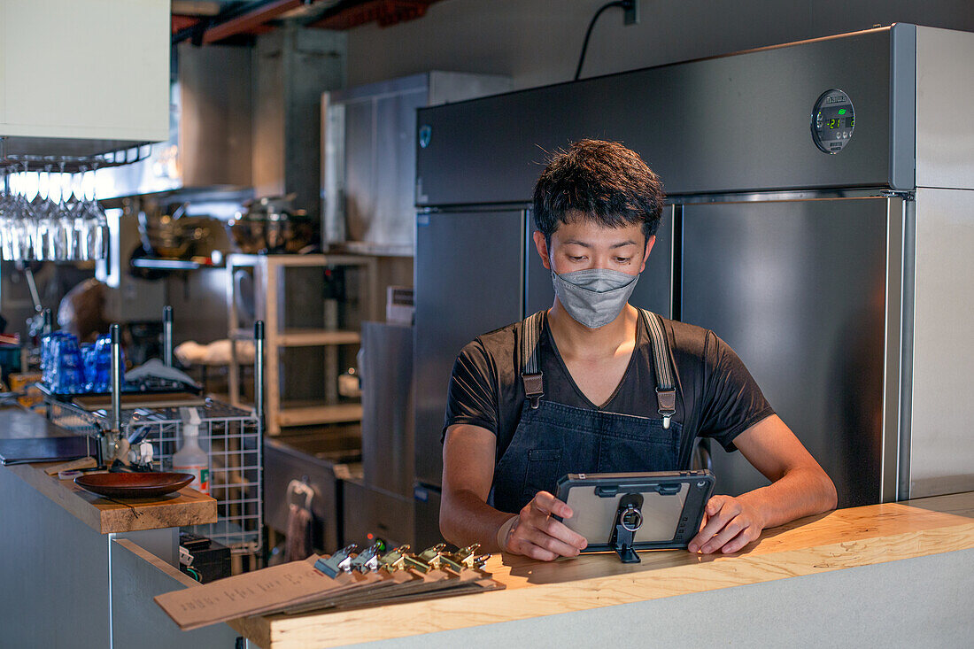 Ein Mann mit Gesichtsmaske an der Theke einer Restaurantküche, der ein digitales Tablet benutzt, der Eigentümer oder Manager. 