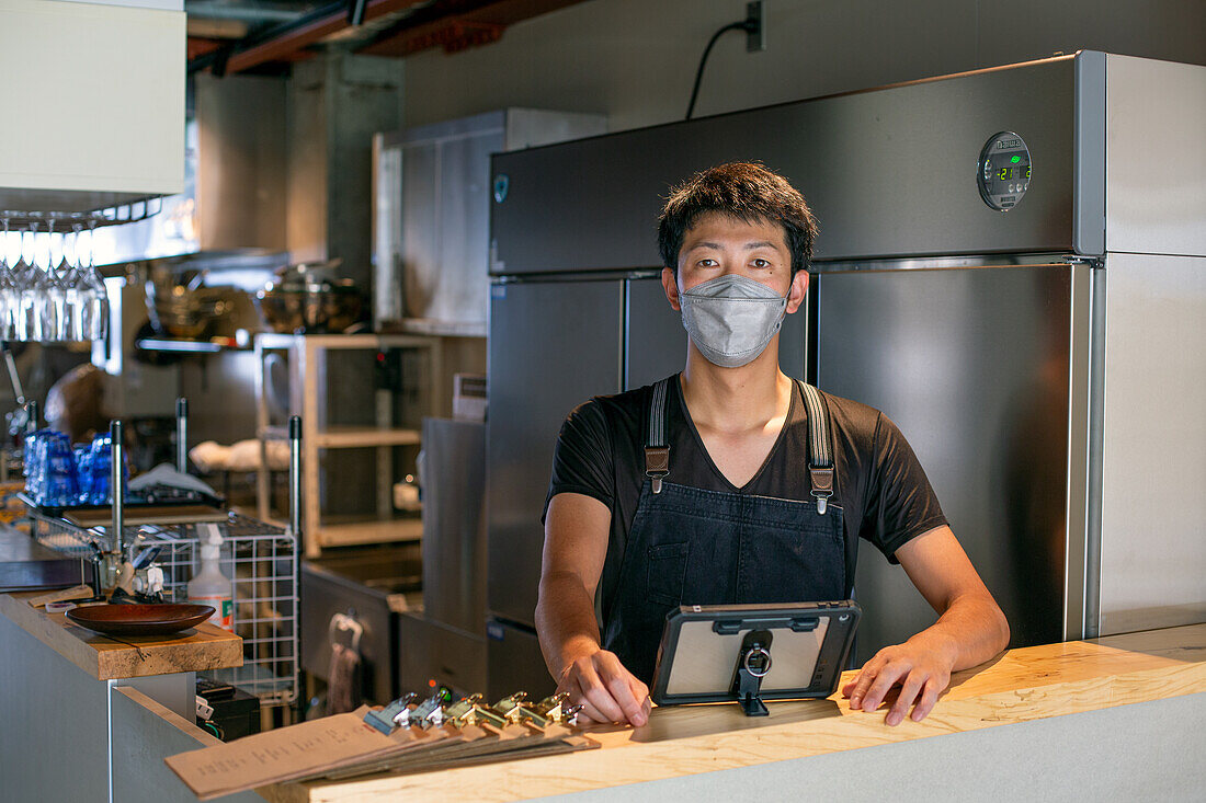 A man wearing a face mask at the counter of a restaurant kitchen, using a digital tablet, the owner or manager. 