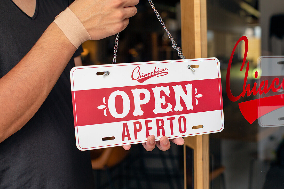 Ein Mann hängt ein Schild "Open" an eine Restauranttür. Zweisprachig, italienisch und englisch. 