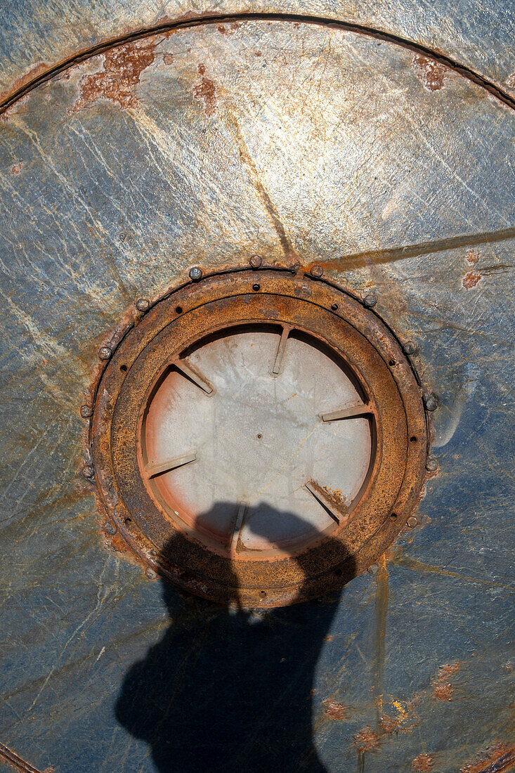 The shadow of a person with arms raised thrown onto the rusted marked surface of a large container with round opening. 