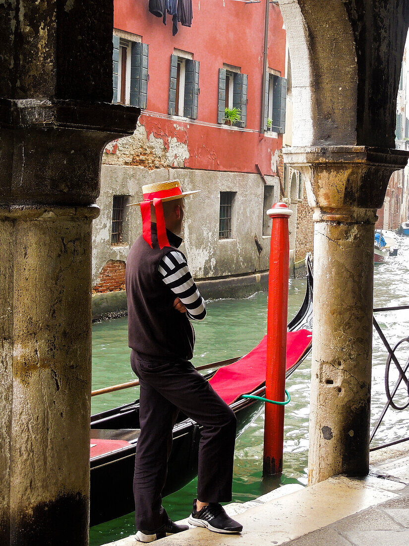 Venedig, Italien. Gondoliere schaut neben seiner Gondel auf den Kanal hinaus