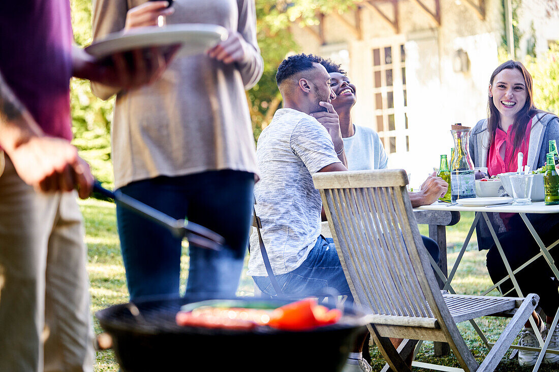 Junger Mann bereitet Essen auf dem Grill zu, während seine Freunde im Hintergrund am Tisch sitzen