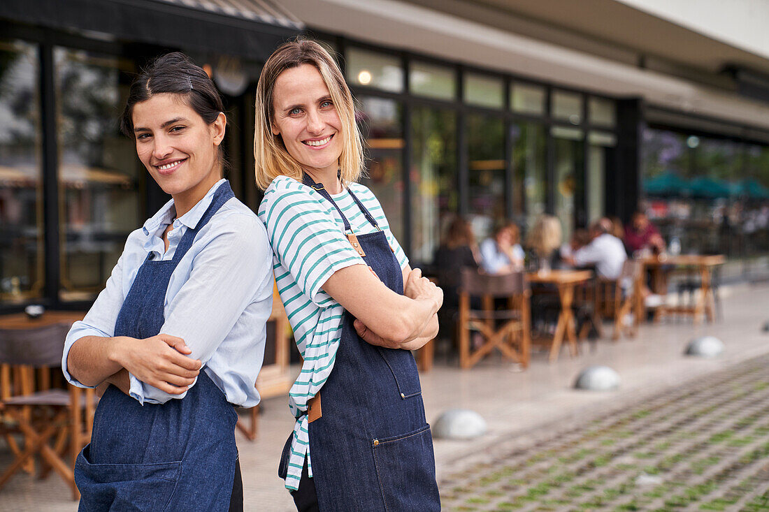Foto von zwei glücklichen Restaurantbesitzerinnen, die Rücken an Rücken vor ihrem Restaurant stehen