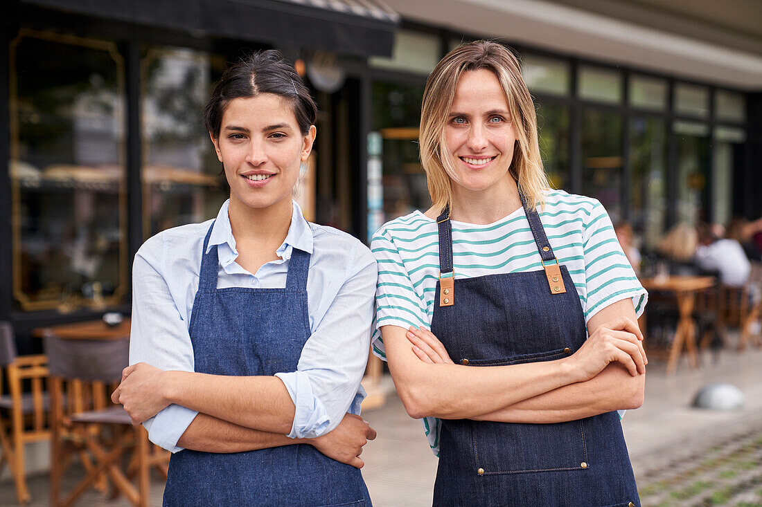Mid shot of two female restaurant entrepreneurs posing in front of their store