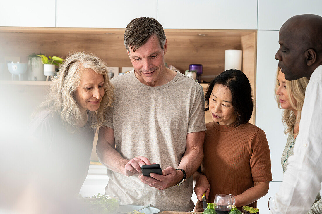 Diverse group of senior friends stading in the kitchen while checking recipe on cell phone