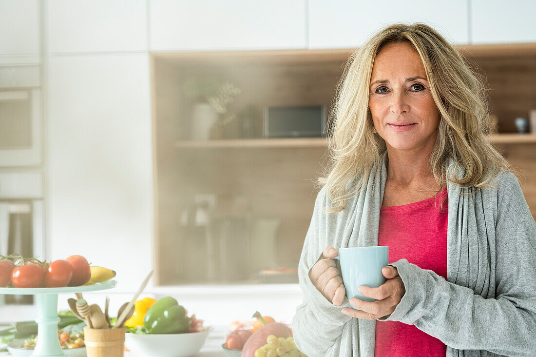 Porträt einer Frau mittleren Alters, die eine Tasse Kaffee hält und in die Kamera schaut, während sie in der Küche steht