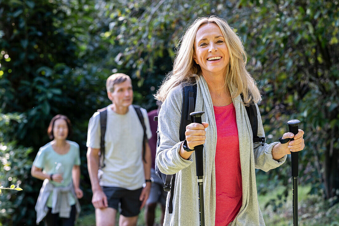 Blonde Dame mittleren Alters mit Rucksack und Wanderstöcken beim Wandern im Wald mit einer Gruppe von Freunden