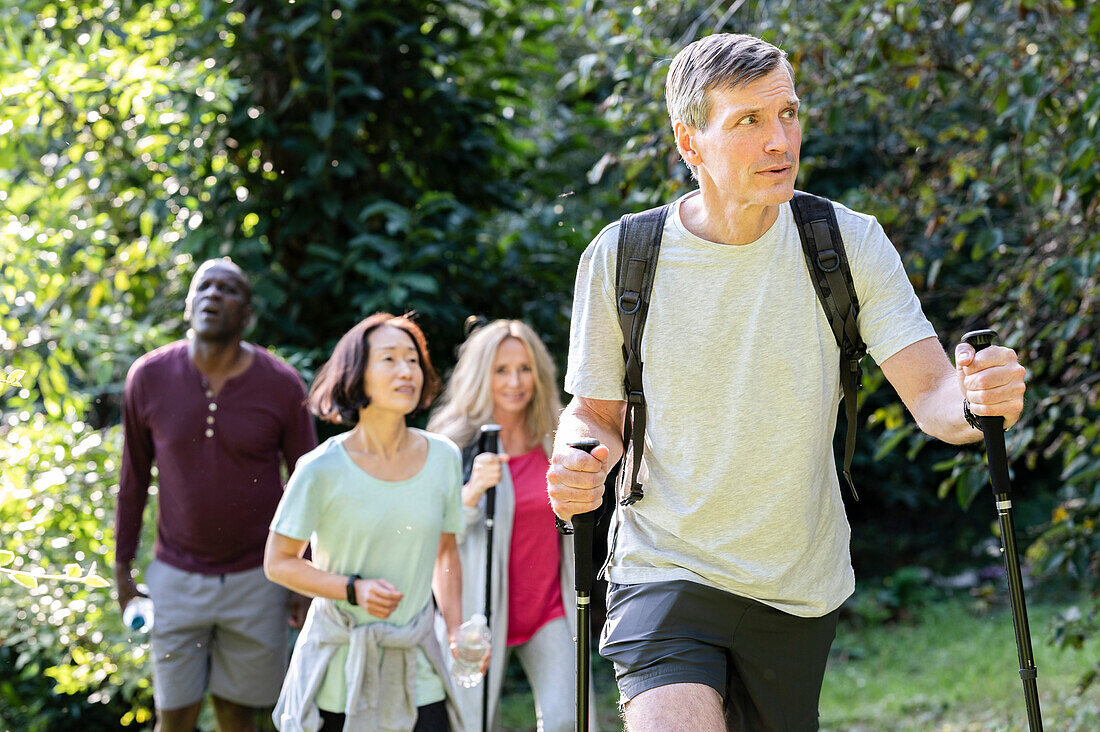 Mann mittleren Alters mit Rucksack und Wanderstöcken beim Wandern im Wald mit einer Gruppe von Freunden