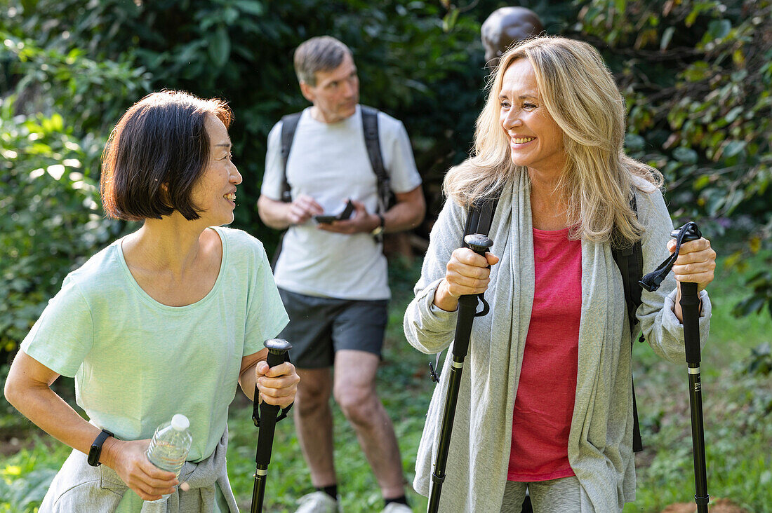 Zwei Damen mittleren Alters mit Wanderstöcken wandern im Wald mit ihren Partnern hinter ihnen