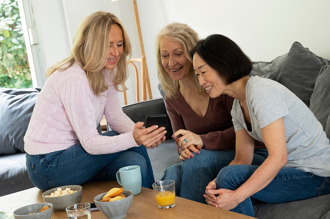 Porträt von drei Frauen mittleren Alters, die Spaß haben, während sie sich im Wohnzimmer Fotos auf dem Handy ansehen