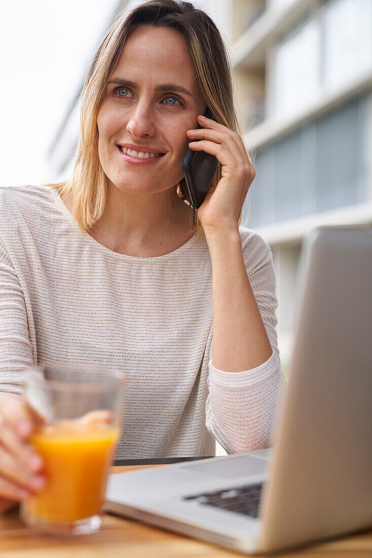 Frontalaufnahme einer Frau, die vor einem Laptop Orangensaft trinkt und mit einem Handy telefoniert