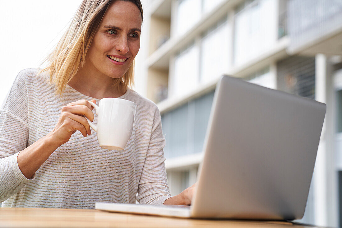 Low angle view of female entrpreneur looking at laptop screen and drinking coffee