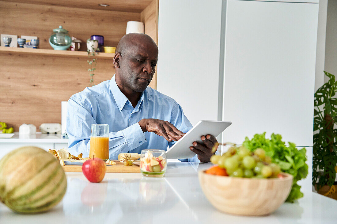 Senior African-American man sitting in kitchen eating fruit for breakfast while checking messages on digital tablet