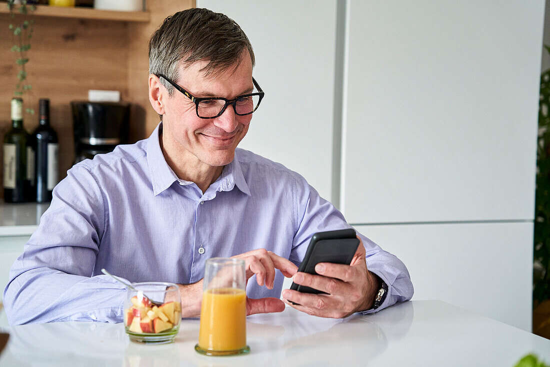 Berufstätiger Mann mittleren Alters, der lächelt und Nachrichten auf dem Telefon abruft, während er zu Hause an der Küchentheke frühstückt