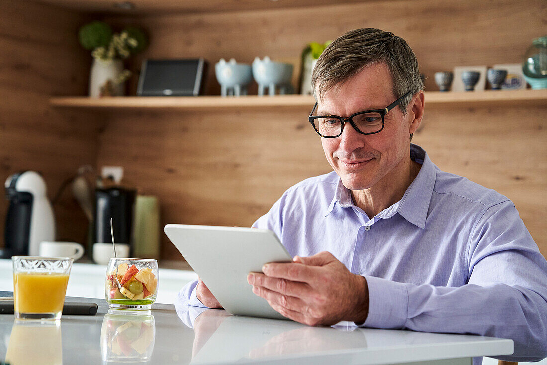 Berufstätiger Mann mittleren Alters, der lächelt und Nachrichten auf einem digitalen Tablet abruft, während er zu Hause an der Küchentheke frühstückt