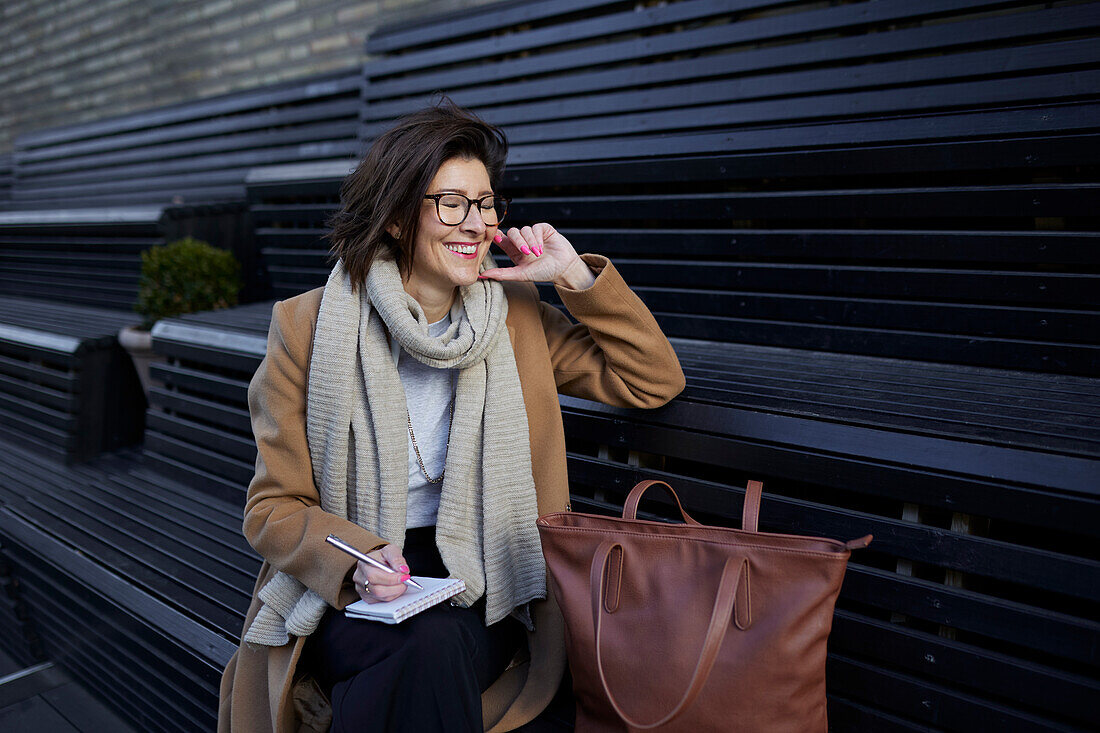 Smiling businesswoman sitting on bench and writing in notebook