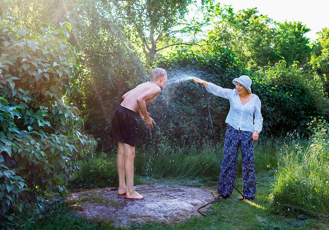 Frau im Garten besprüht Mann mit Gartenschlauch