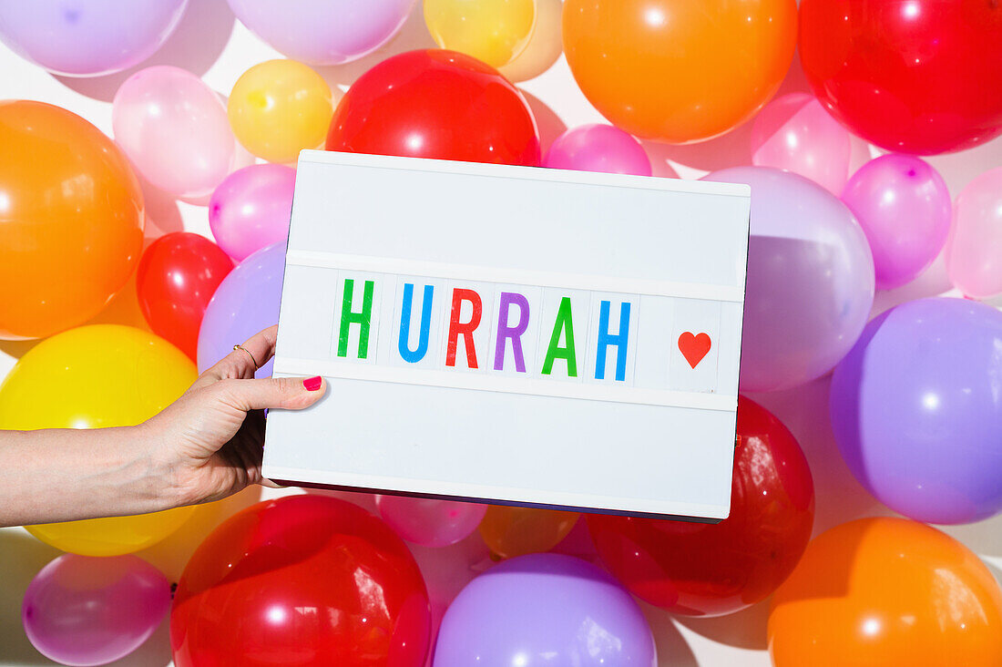 Frau hält Schild mit der Aufschrift "Hurra" vor einer Luftballonwand