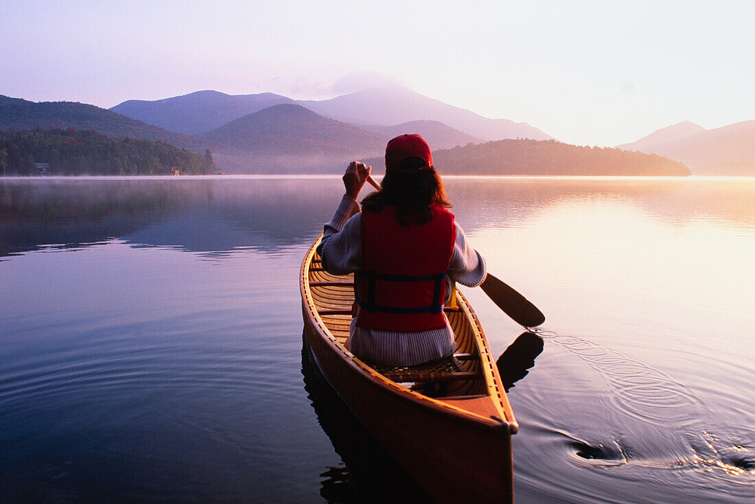 United States, New York, Rear view of woman paddling canoe on Lake Placid at sunrise, Adirondacks State Park