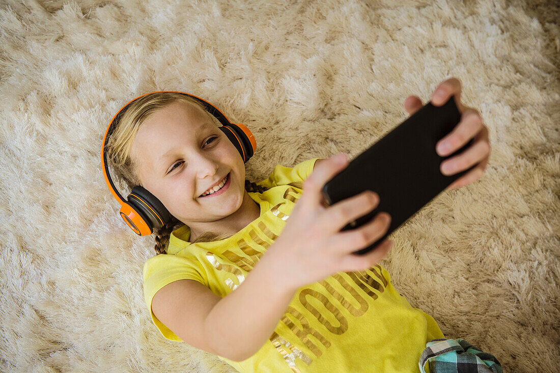 Smiling girl (10-11) with headphones and smart phone lying on hairy rug