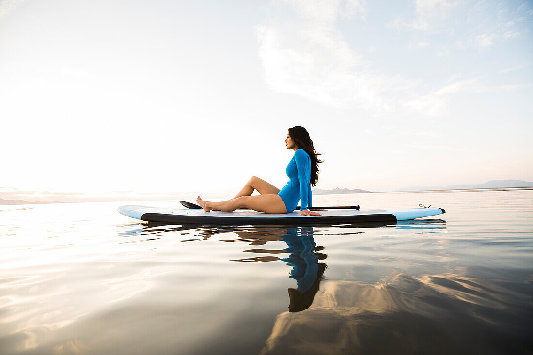 Porträt einer Frau in blauem Badeanzug auf einem Paddleboard auf einem See bei Sonnenuntergang sitzend